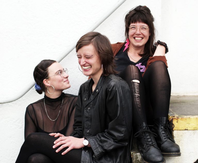 Die Band Teddies Kneipe, bestehend aus ihren drei weiblichen Mitgliedern sitzen nebeneinander auf einer Treppe und lachen sich gegenseitig an.