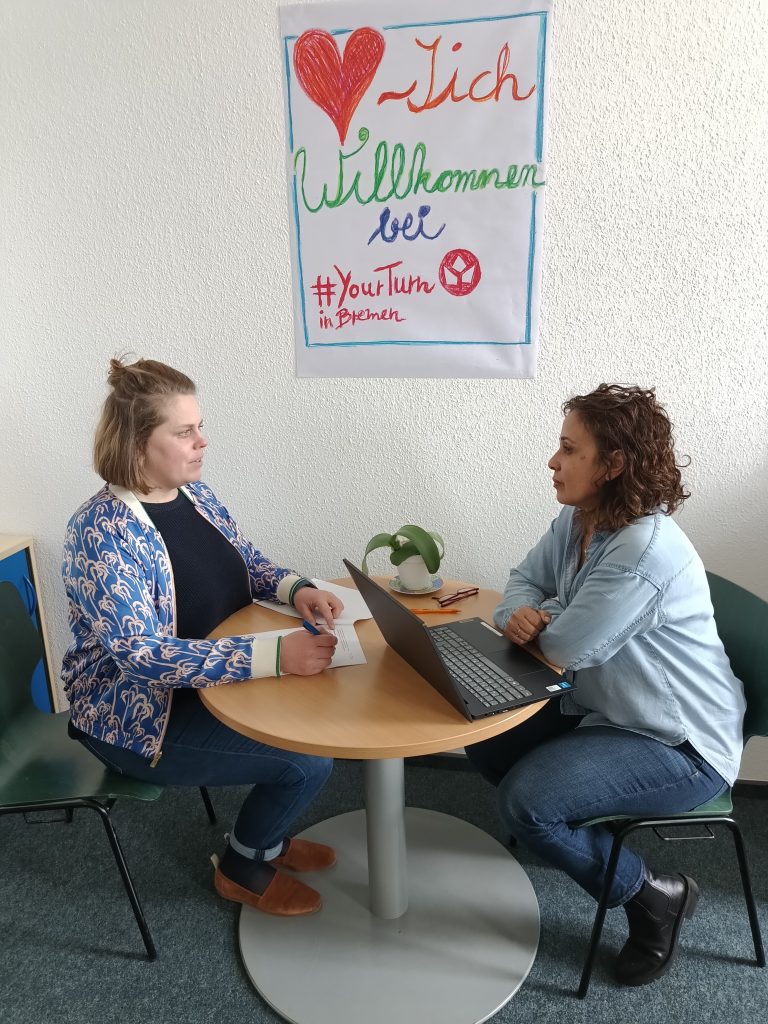 Zwei Frauen sitzen an einem Tisch und unterhalten sich. Eine hat einen Laptop vor sich, die andere schreibt auf ein Papier. An der Wand hängt ein Plakat mit der Aufschrift "Herzlich Willkommen bei #YourTurn in Bremen".