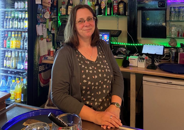 Das Bild zeigt die Inhaberin der Bar Oililio. Sie steht hinter der Bar und lächelt in die Kamera.