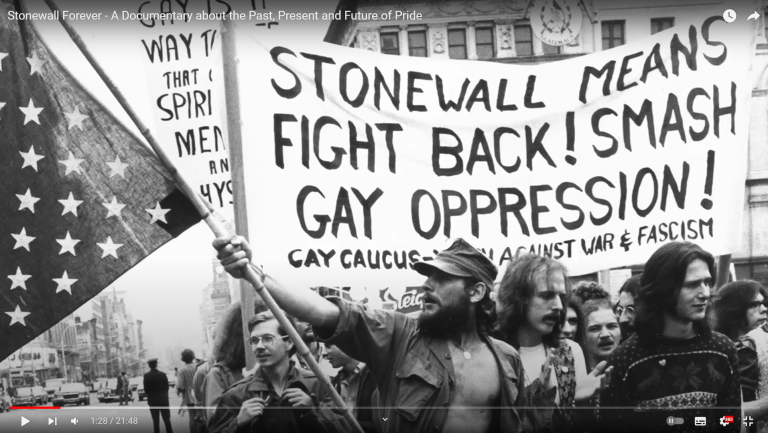 Auf dem Bild sind mehrere demonstrierende Menschen zu sehen und eine Flagge mit der Aufschrift "Stonewall means fight back! Smash gay opression!"