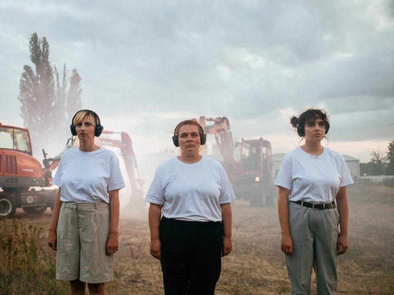 Drei Frauen stehen in einer Reihe auf einem Brachgelände. Hinter ihnen ist Staub und Nebel aus dem zwei Bagger hervortauchen.