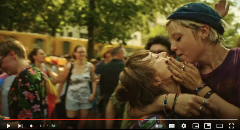 Ein Screenshot aus dem Trailer zu dem Film "Kokon". Man sieht wie die Charaktere Nora und Romy sich in den Armen halten. Sie sind auf einem Straßenfest, hinter ihnen sind andere Menschen und das Bild hat eine sommerliche, fröhliche Stimmung.