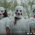 Man sieht einen Screenshot von dem Musikvideo "SWAN LAKE" von Pussy Riot. Zu sehen sind 3 Mitgliederinnen mit weißen Sturmmasken auf denen rote Kreise als Bäckchen aufgeklebt sind. Hinter ihnen ist ein Wald zu sehen.