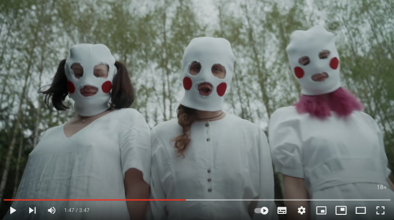 Man sieht einen Screenshot von dem Musikvideo "SWAN LAKE" von Pussy Riot. Zu sehen sind 3 Mitgliederinnen mit weißen Sturmmasken auf denen rote Kreise als Bäckchen aufgeklebt sind. Hinter ihnen ist ein Wald zu sehen.