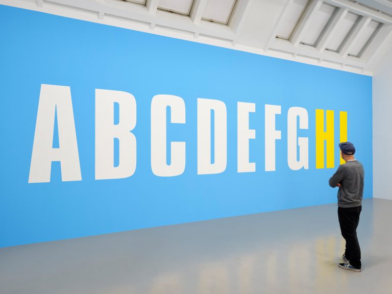 Kunstwerk "HI" von Kay Rosen mit Museumsbesucher*in zu sehen sind die Buchstaben A B C D E F G in weißer Farbe und H und I in gelber Farbe