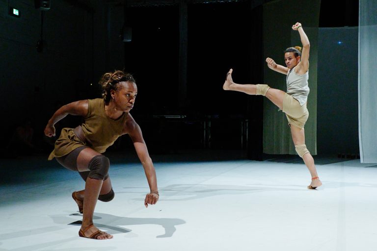 Man sieht einen Ausschnitt der Tanzperformance Azúcar von Antonya Silva. Die Tänzerin Antonya Silva steht in schleichender Position. Ein anderer Tänzer im Hintergrund wirft sein Bein in die Luft.