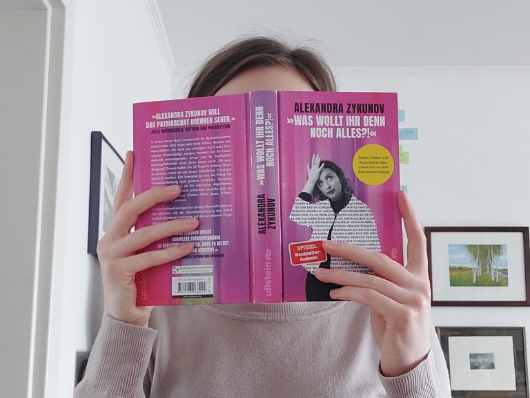 Eine junge Frau hält ein Buch vor ihr Gesicht, das Cover ist pink und die Autorin ist darauf abgebildet.