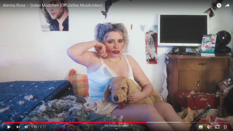 Man sieht einen Screenshot von einem Youtube-Video. Darauf erkennt man eine Frau, die auf ihrem Bett sitzt. Das Zimmer ist unordentlich. Sie hat ein Kuscheltier auf dem Schoß und wischt sich ihren Lippenstift mit der Hand von den Lippen runter. Im Hintergrund sieht man Poster und einen Bildschirm.