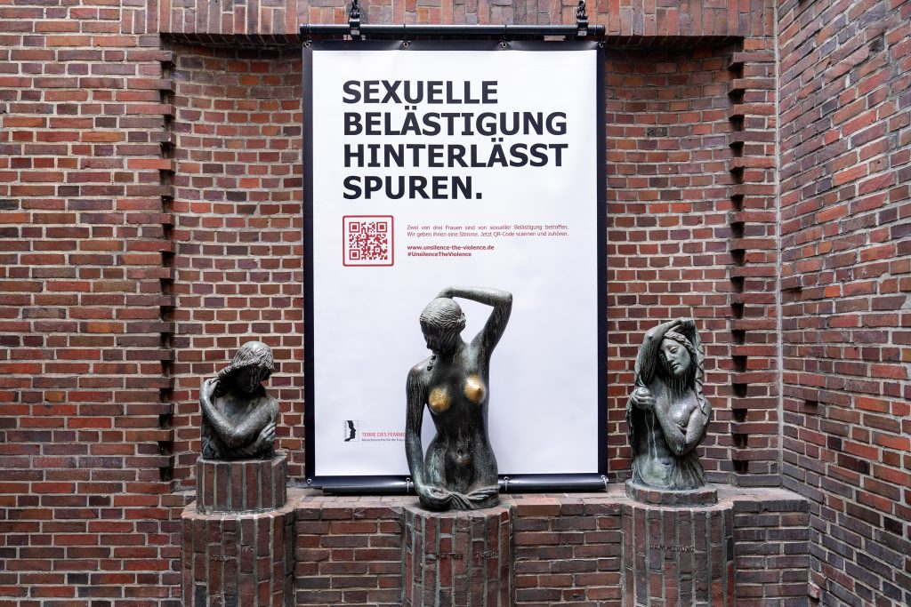 Drei weibliche Bronzestatuen, die in der Mitte hat durch häufiges Betatschen helle Brüste. Hinter der Statue hängt ein großes Plakat mit der Aufschrift "Sexuelle Belästigung hinterlässt Spuren"