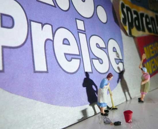 Miniatur-Hausfrau steht  vor Wand mit Spruch "k.o. Preise"